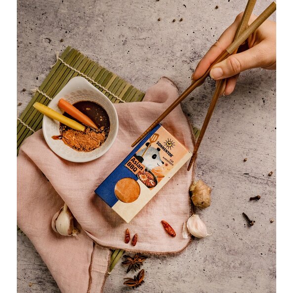 Asian BBQ Gewürz Packung neben Schale mit Marinade, Ingwer, Knoblauch und Essstäbchen auf hellem asiatisch dekoriertem Untergrund.